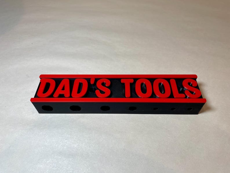 May include: A black tool organizer with the text "DAD'S TOOLS" in red. The organizer has a red border and several holes for holding tools. The item is made of plastic and is designed to hold tools.