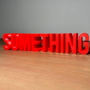 Pu&ograve; includere: La parola "SOMETHING" in rosso vivo, tridimensionale, &egrave; esposta su una superficie di legno. Le lettere a blocchi sono spaziate uniformemente, proiettando ombre. Lo sfondo &egrave; grigio neutro.