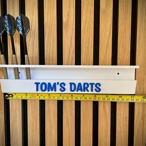 May include: White dart holder with the text "TOM'S DARTS" in blue. Three darts are in the holder. A yellow tape measure is below the holder. The background is a wooden wall.