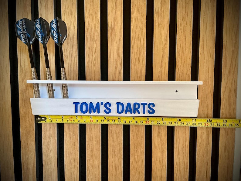 May include: White dart holder with the text "TOM'S DARTS" in blue. Three darts are in the holder. A yellow tape measure is below the holder. The background is a wooden wall.