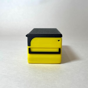 Pu&ograve; includere: Un piccolo contenitore rettangolare giallo con un coperchio nero e dettagli neri. Il contenitore ha un piccolo foro rotondo sul lato. Il design &egrave; moderno e minimalista.
