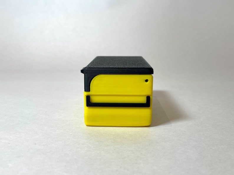 Pu&ograve; includere: Un piccolo contenitore rettangolare giallo con un coperchio nero e dettagli neri. Il contenitore ha un piccolo foro rotondo sul lato. Il design &egrave; moderno e minimalista.