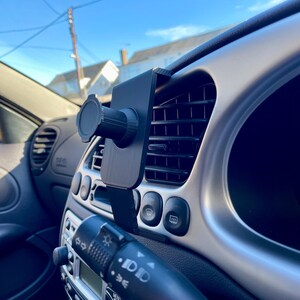May include: A black car phone mount attached to the air vent in a car's dashboard. The mount has a ball joint for adjusting the phone's angle.