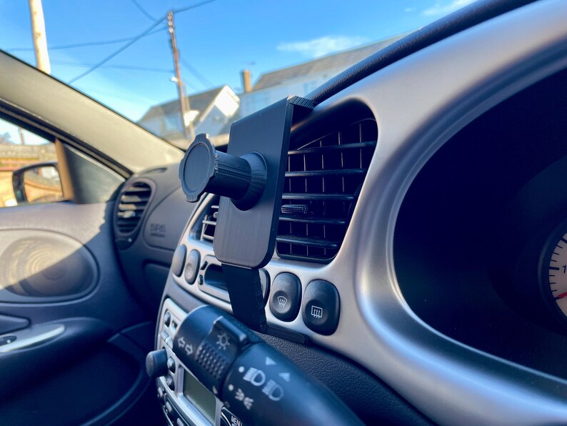 May include: A black car phone mount attached to the air vent in a car's dashboard. The mount has a ball joint for adjusting the phone's angle.