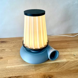 Puede incluir: Una l&aacute;mpara de mesa azul y blanca con una pantalla de tela plisada. La l&aacute;mpara tiene una base &uacute;nica que se asemeja a un ventilador.