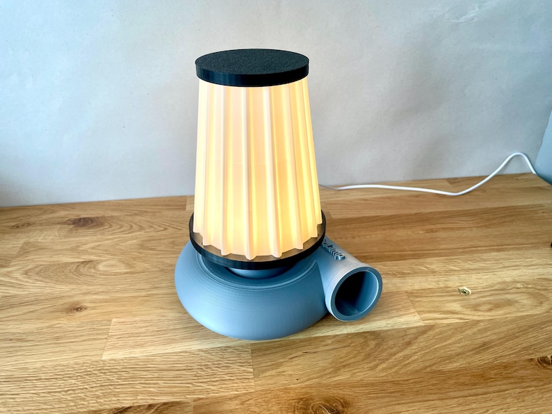 Puede incluir: Una l&aacute;mpara de mesa azul y blanca con una pantalla de tela plisada. La l&aacute;mpara tiene una base &uacute;nica que se asemeja a un ventilador.