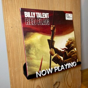 May include: A vinyl record by Billy Talent titled "Red Flag" is displayed on a wooden record holder with a black "NOW PLAYING" sign. The album cover features a person holding a red and white flag.