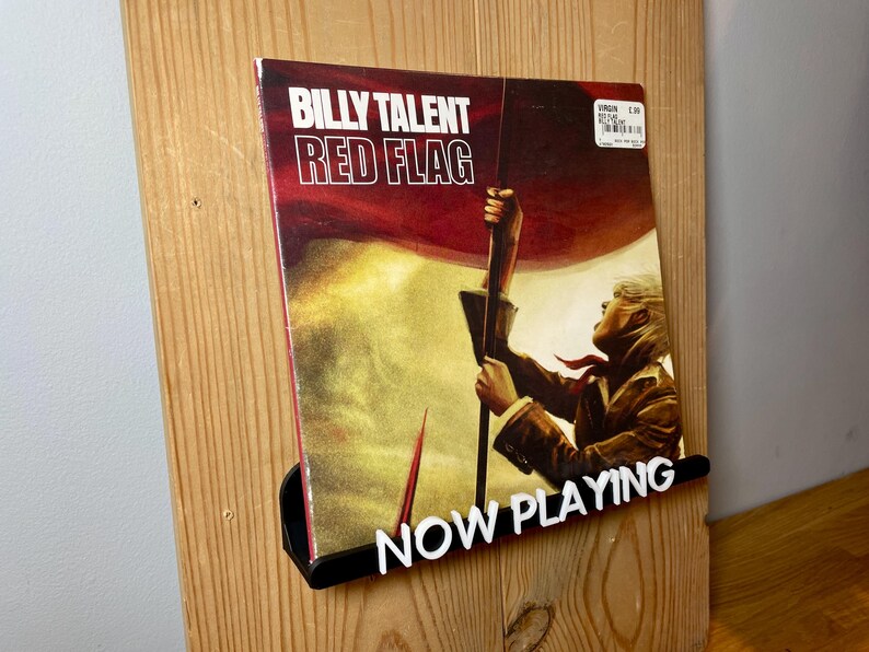 May include: A vinyl record by Billy Talent titled "Red Flag" is displayed on a wooden record holder with a black "NOW PLAYING" sign. The album cover features a person holding a red and white flag.