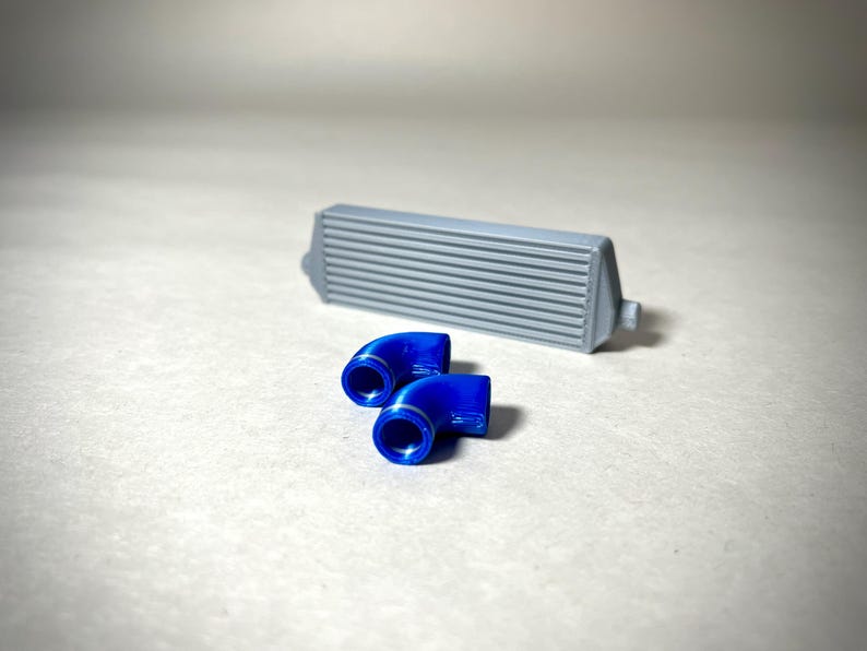 May include: A gray intercooler with horizontal fins and two bright blue, curved pipe fittings. The components are likely for an automotive or mechanical application, set against a neutral background.