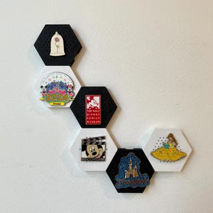 May include: A collection of Disney-themed pins displayed on hexagonal black and white magnetic boards. The pins feature characters and logos from Disney films and parks, including Mickey Mouse, Belle, and Disneyland.
