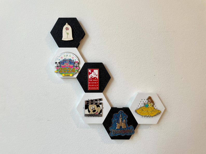 May include: A collection of Disney-themed pins displayed on hexagonal black and white magnetic boards. The pins feature characters and logos from Disney films and parks, including Mickey Mouse, Belle, and Disneyland.