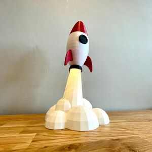 Op de afbeelding: Een witte en rode raketlamp met een zwart patrijspoort en rode vinnen. De raket straalt een geel licht uit vanaf de basis, met witte rookachtige wolken. De lamp staat op een houten oppervlak.