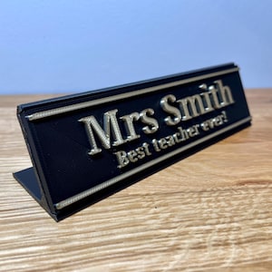 name plaque / plate with a teachers name and best teacher ever! text on the front. Black with bronze text on a wooden desk with grey background