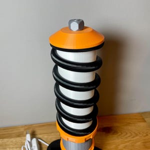 Op de afbeelding: Een 3D-geprinte oranje en zwarte tafellamp in de vorm van een autoveer. De lamp heeft een witte lichtbron en een zilveren bovenkant.