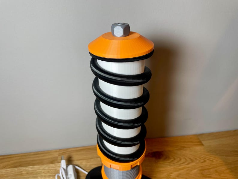 Op de afbeelding: Een 3D-geprinte oranje en zwarte tafellamp in de vorm van een autoveer. De lamp heeft een witte lichtbron en een zilveren bovenkant.
