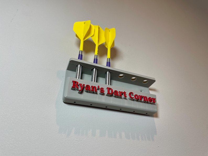 Wall mounted dart holder. Grey with red personalised text on the front reading Ryan's Dart Corner