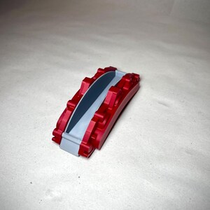 May include: A small, red and gray decorative bridge. The bridge has a curved design with a gray pathway and red supports. The object is made of a solid material and sits on a neutral surface.