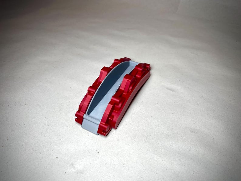 May include: A small, red and gray decorative bridge. The bridge has a curved design with a gray pathway and red supports. The object is made of a solid material and sits on a neutral surface.