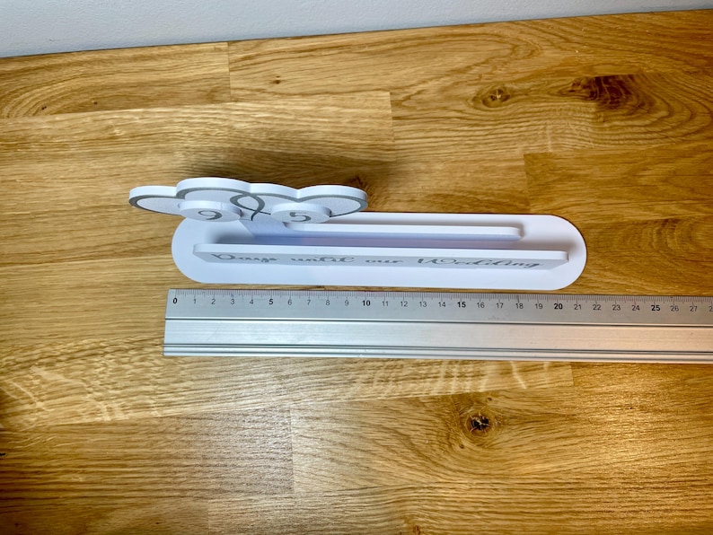 May include: White wooden sign with the text "Days until our wedding" and a number display. The sign is sitting on a wooden surface with a ruler next to it.