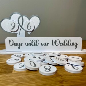 May include: White wooden countdown sign with two silver glitter hearts and the text "Days until our Wedding". The sign has a set of white numbered circles for counting down the days.