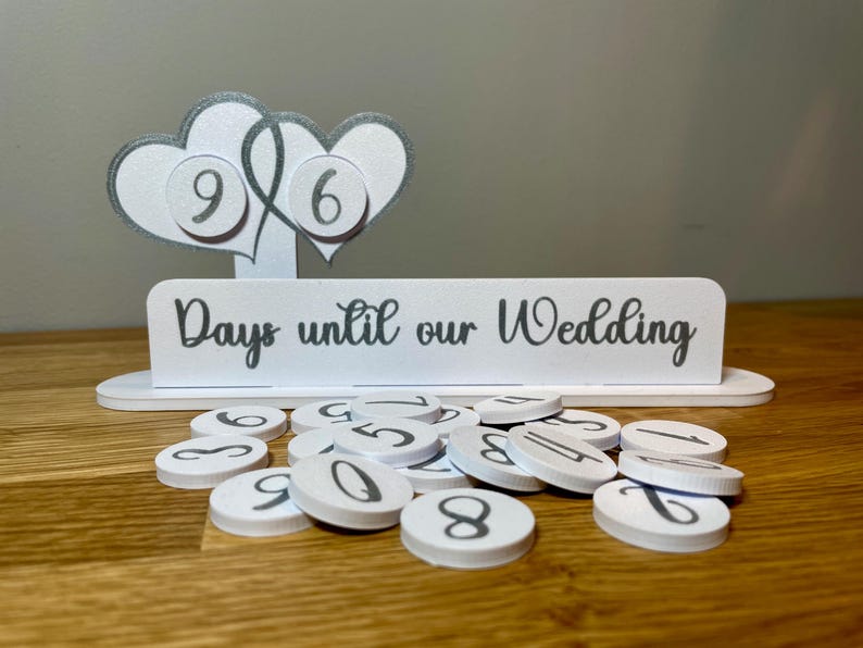 May include: White wooden countdown sign with two silver glitter hearts and the text "Days until our Wedding". The sign has a set of white numbered circles for counting down the days.