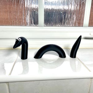 May include: A black, 3-piece, 3D printed, cartoon-style, worm or snake with googly eyes. The worm is segmented and has a curved body.