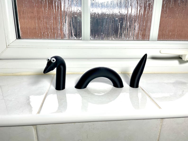 May include: A black, 3-piece, 3D printed, cartoon-style, worm or snake with googly eyes. The worm is segmented and has a curved body.