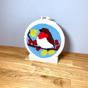 May include: A white embroidery hoop with a blue background and a red-breasted robin perched on a branch with yellow leaves. The robin is white, red, and brown. The hoop is on a wooden stand.