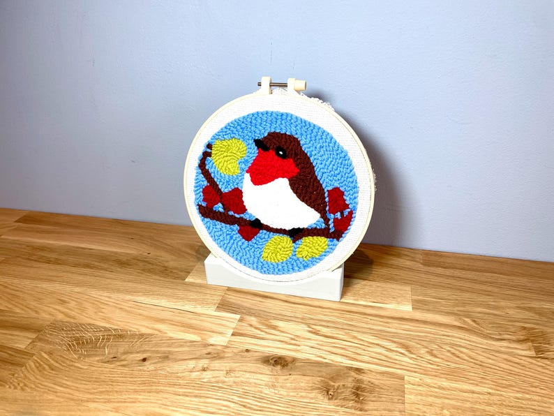 May include: A white embroidery hoop with a blue background and a red-breasted robin perched on a branch with yellow leaves. The robin is white, red, and brown. The hoop is on a wooden stand.