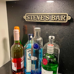 May include: A black countertop with a gold sign that reads "STEVE'S BAR". There are five bottles on the counter, including a pink wine bottle, a clear bottle with a blue label, a clear bottle with a green label, and two clear bottles with blue labels.