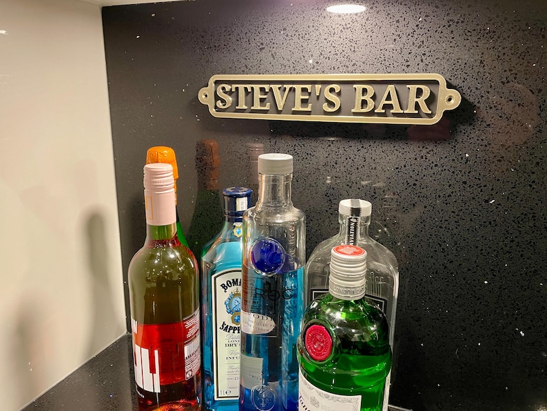 May include: A black countertop with a gold sign that reads "STEVE'S BAR". There are five bottles on the counter, including a pink wine bottle, a clear bottle with a blue label, a clear bottle with a green label, and two clear bottles with blue labels.