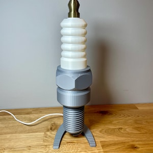 Oversized spark plug lamp with the white ceramic section designed as a light shade. Bronze top section, white shade and silver base. With a white cable. Stood on a wooden desk and grey background.
