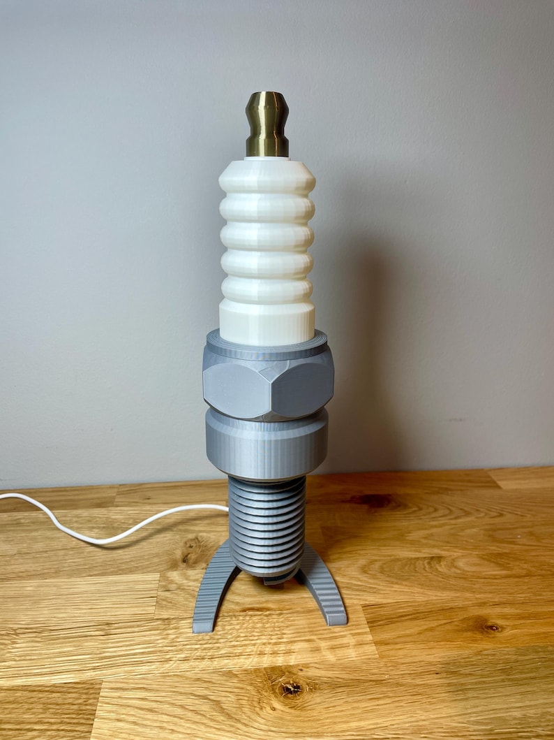 Oversized spark plug lamp with the white ceramic section designed as a light shade. Bronze top section, white shade and silver base. With a white cable. Stood on a wooden desk and grey background.