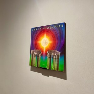 May include: A vintage vinyl record album cover featuring a colorful illustration of a sun with rays of light shining down on a row of columns. The album cover is titled "Earth, Wind & Fire" in a stylized font.