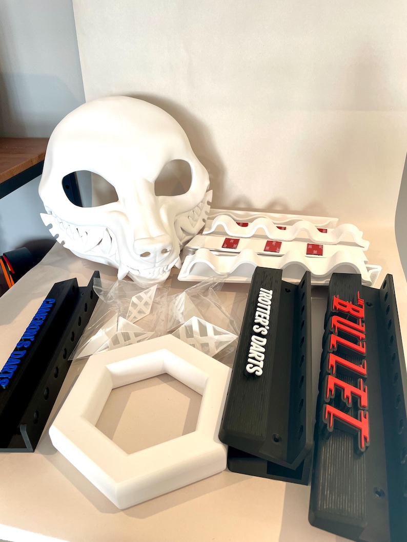 May include: A collection of white and black 3D-printed items. Includes a white animal mask, black dart holders with "TROTTER'S DARTS" and "BULLET" text, and a white hexagonal frame. Also, there are some white plastic pieces.