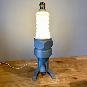 Oversized spark plug lamp with the white ceramic section designed as a light shade. Bronze top section, white shade and silver base. With a white cable. Stood on a wooden desk and grey background.