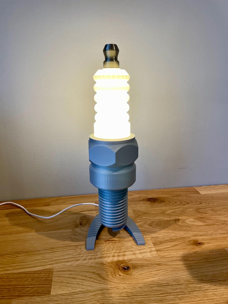 Oversized spark plug lamp with the white ceramic section designed as a light shade. Bronze top section, white shade and silver base. With a white cable. Stood on a wooden desk and grey background.