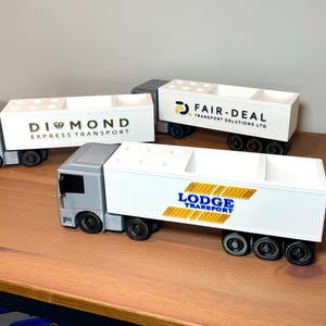 Pu&ograve; includere: Tre camion giocattolo con rimorchi bianchi e cabine grigie sono esposti su una superficie di legno. Ogni rimorchio ha un logo aziendale diverso: Diamond Express Transport, Fair-Deal Transport Solutions LTD e Lodge Transport.