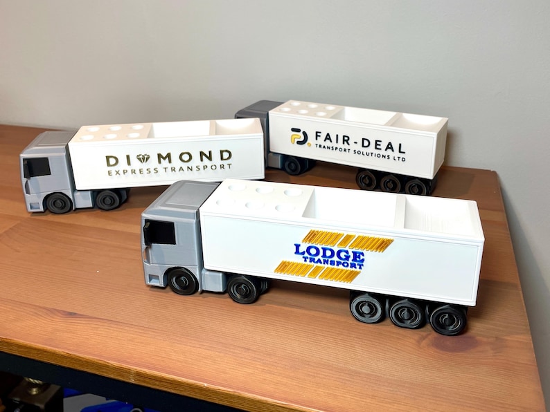 Pu&ograve; includere: Tre camion giocattolo con rimorchi bianchi e cabine grigie sono esposti su una superficie di legno. Ogni rimorchio ha un logo aziendale diverso: Diamond Express Transport, Fair-Deal Transport Solutions LTD e Lodge Transport.