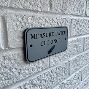 May include: A rectangular gray sign with black text that reads "MEASURE TWICE CUT ONCE" and a saw graphic. The sign is mounted on a white brick wall. The sign has a black border.