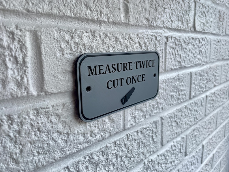 May include: A rectangular gray sign with black text that reads "MEASURE TWICE CUT ONCE" and a saw graphic. The sign is mounted on a white brick wall. The sign has a black border.