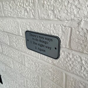 May include: A rectangular, silver-colored sign with rounded corners is mounted on a white brick wall. The sign features the text "There's two ways to do things: 1. The right way 2. Again" in black lettering. The wall has a textured surface.