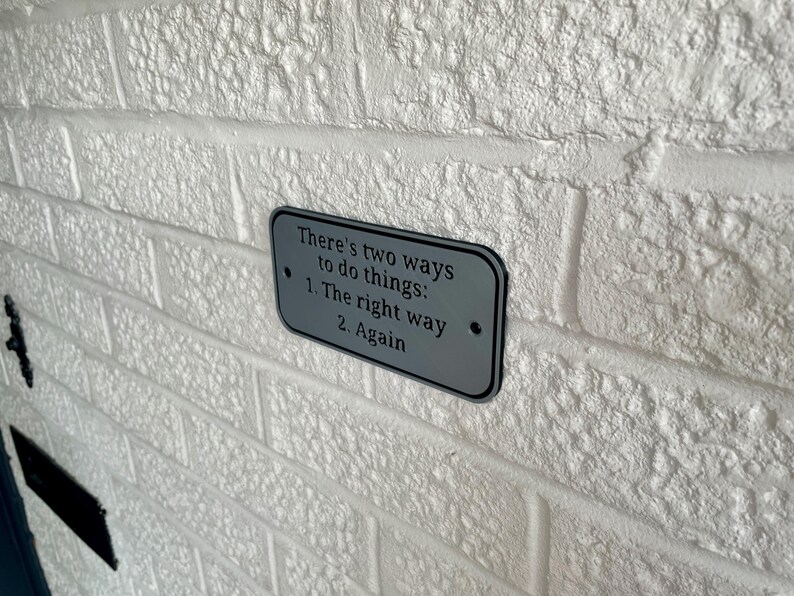 May include: A rectangular, silver-colored sign with rounded corners is mounted on a white brick wall. The sign features the text "There's two ways to do things: 1. The right way 2. Again" in black lettering. The wall has a textured surface.