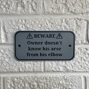 Puede incluir: Un letrero rectangular gris con esquinas redondeadas est&aacute; montado en una pared de ladrillos blancos. El letrero dice "BEWARE" encima del texto "Owner doesn't know his arse from his elbow."