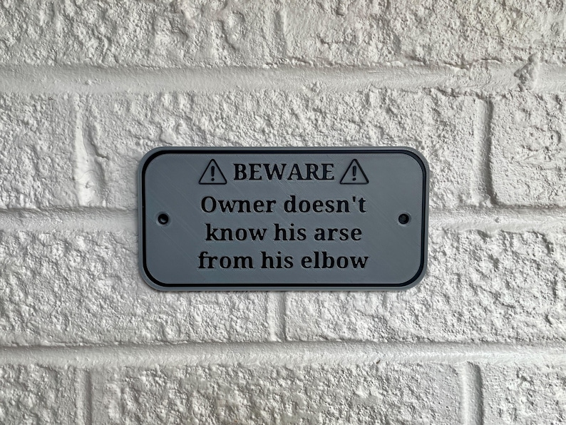 Puede incluir: Un letrero rectangular gris con esquinas redondeadas est&aacute; montado en una pared de ladrillos blancos. El letrero dice "BEWARE" encima del texto "Owner doesn't know his arse from his elbow."