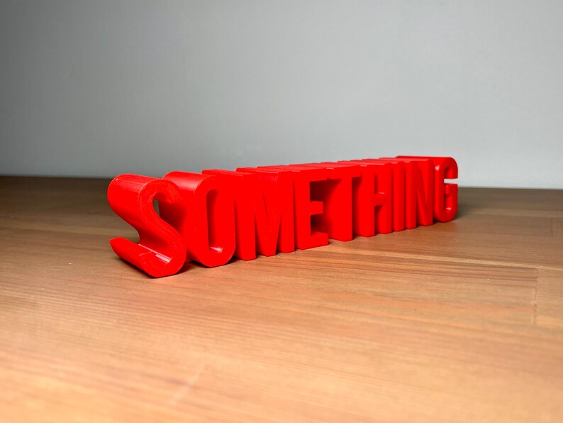 Pu&ograve; includere: Un cartello rosso brillante, stampato in 3D, con la scritta "SOMETHING". Le lettere a blocchi sono su una superficie di legno. Un elemento decorativo accattivante.