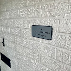 Puede incluir: Un letrero rectangular de metal gris con esquinas redondeadas est&aacute; montado en una pared de ladrillos blancos. El letrero presenta la cita: "Si al principio no tienes &eacute;xito, int&eacute;ntalo, int&eacute;ntalo de nuevo". La pared tiene una superficie texturizada.