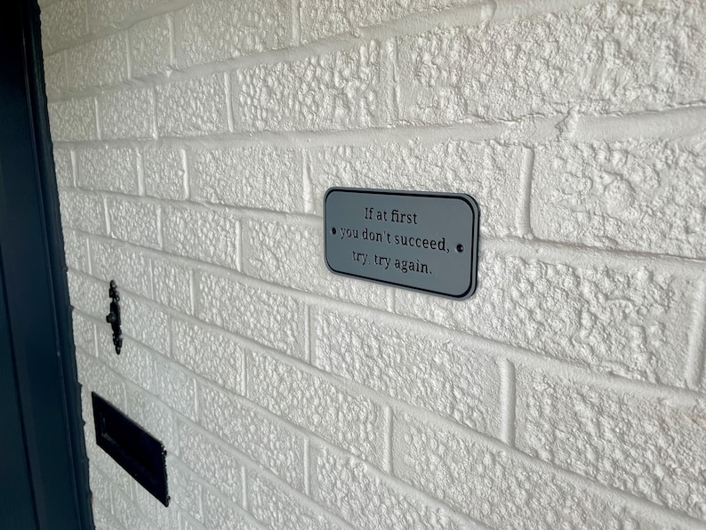 Puede incluir: Un letrero rectangular de metal gris con esquinas redondeadas est&aacute; montado en una pared de ladrillos blancos. El letrero presenta la cita: "Si al principio no tienes &eacute;xito, int&eacute;ntalo, int&eacute;ntalo de nuevo". La pared tiene una superficie texturizada.