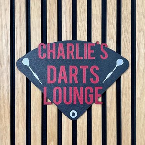 Triangular darts sign with the text charlie's darts lounge in red. Main background is black and has silver inlayed darts logos. On a wooden slatted background.
