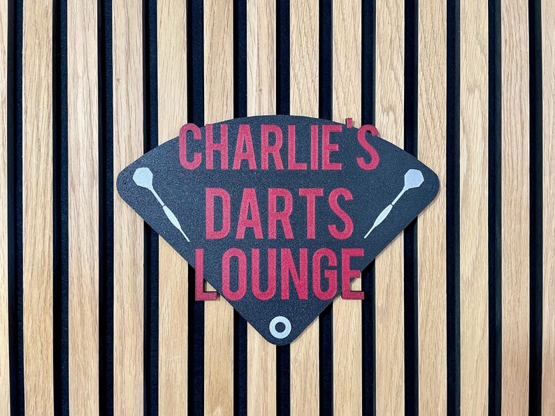 Triangular darts sign with the text charlie's darts lounge in red. Main background is black and has silver inlayed darts logos. On a wooden slatted background.